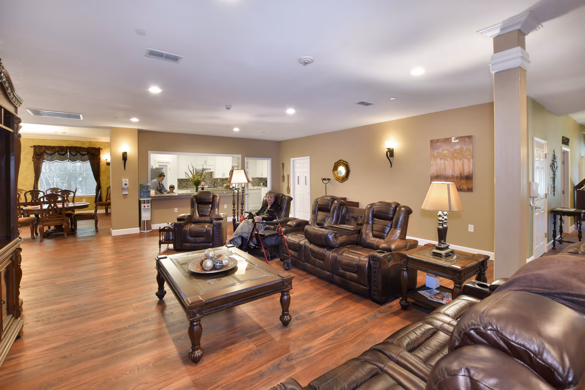 A spacious assisted living facility common area with brown leather recliner sofas and a wooden coffee table. An elderly woman with a walker is seated on one of the recliners. The room has warm beige walls, wooden flooring, and soft lighting from table lamps and wall sconces. In the background, a dining area with a wooden dining table and chairs is visible, along with a kitchen pass-through window where two people are interacting.