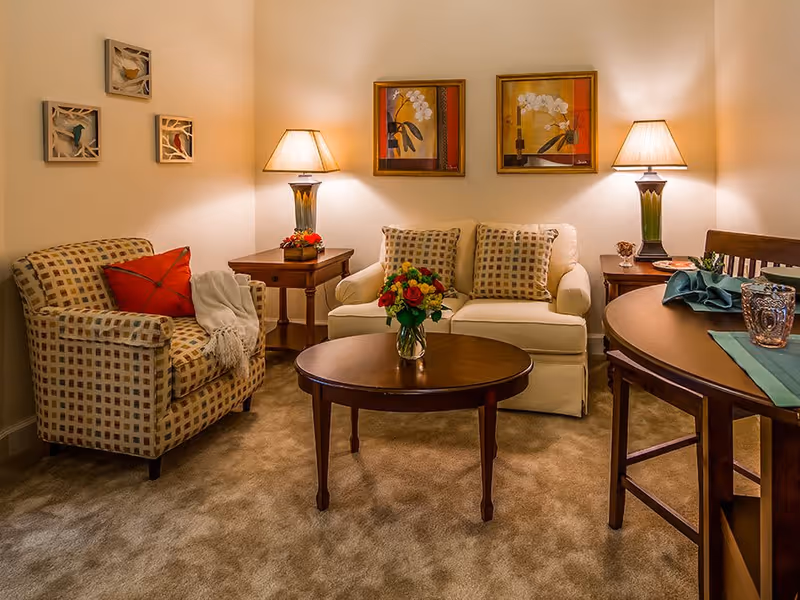 A cozy living room with a beige loveseat and a patterned armchair, both adorned with decorative pillows. A wooden coffee table with a vase of colorful flowers sits in the center. Two table lamps with warm lighting are placed on side tables next to the loveseat. The walls feature framed artwork and small decorative pieces. A round wooden dining table with chairs is partially visible on the right side.