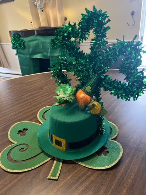 A festive St. Patrick's Day decoration on a wooden table featuring a green top hat with a black band and gold buckle, a small gnome with an orange beard and green hat, and a large green glittery shamrock. In the background, there is a green apron or cloth with shamrock decorations hanging on a chair.