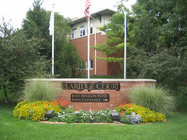 Brick sign for Claridge Court with the address 8101 Mission Road and an arrow pointing to the main entrance, surrounded by green grass, colorful flowers, and tall plants, with a brick building and three flagpoles in the background.