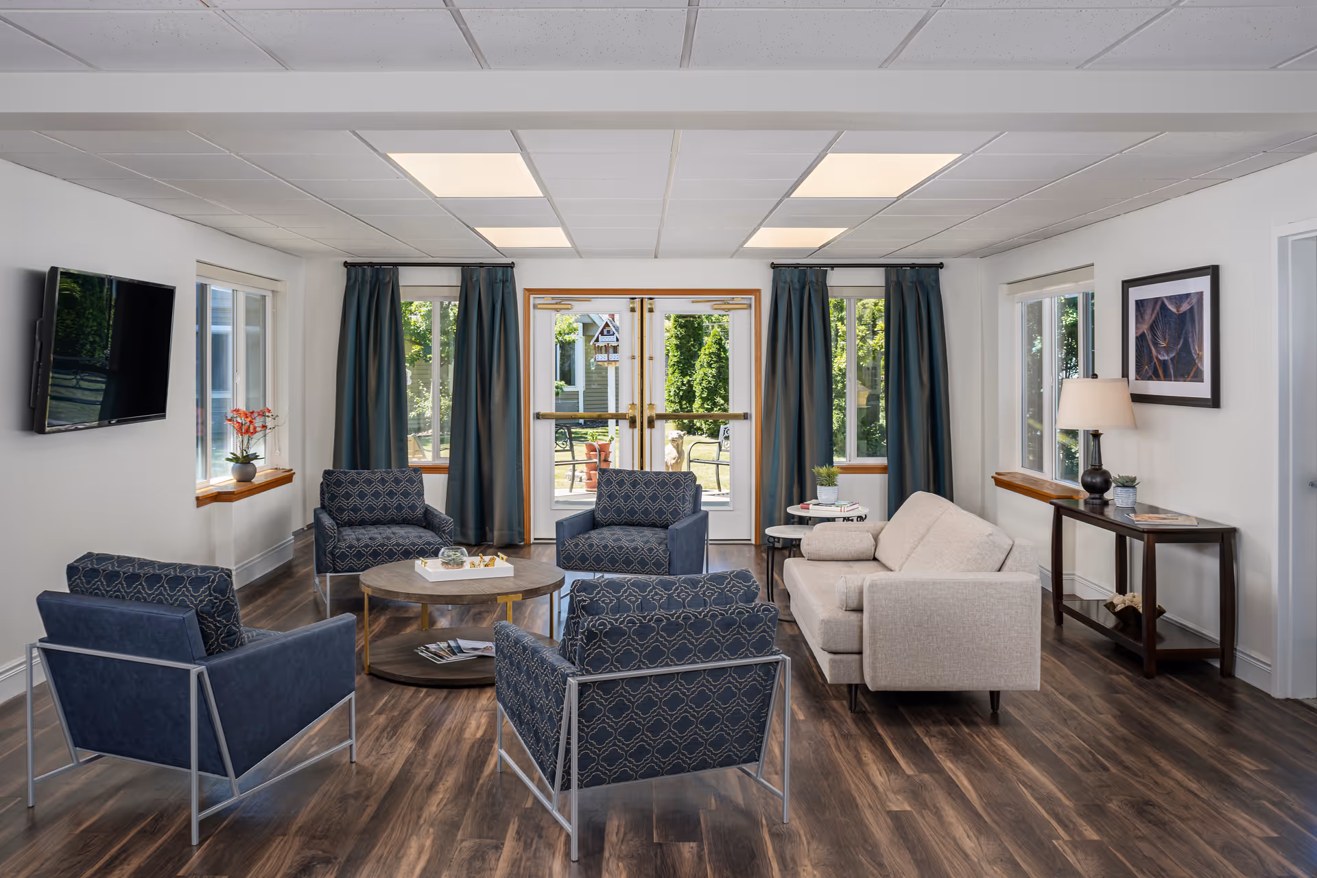 A bright and modern senior living facility common area with four blue patterned armchairs and a beige sofa arranged around a round wooden coffee table. The room has large windows with dark blue curtains, a wall-mounted TV, a console table with a lamp and decorative items, and wood flooring. Outside the glass doors, greenery and outdoor seating are visible.