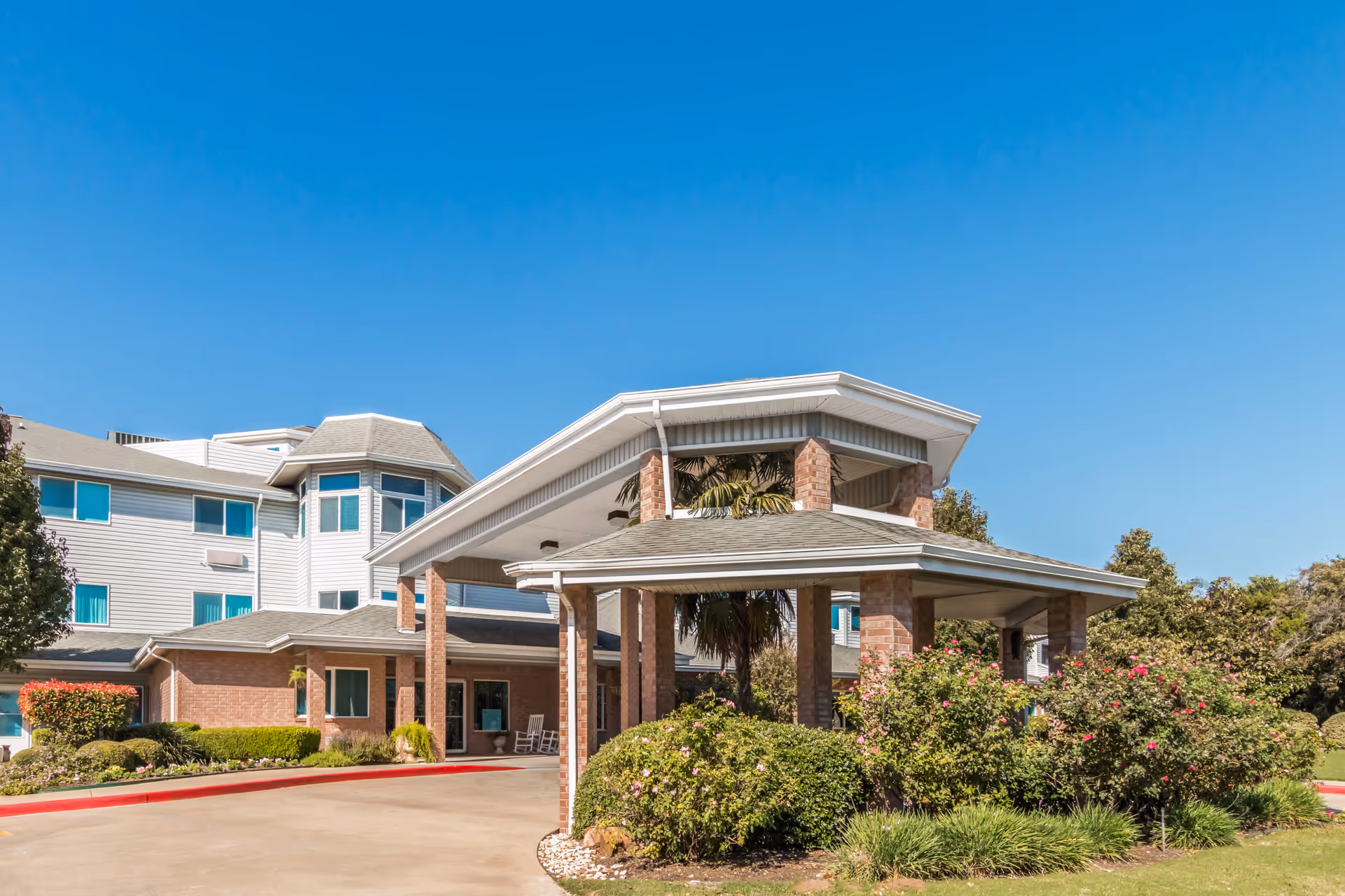Exterior view of a senior living facility building with a covered entrance supported by brick columns, surrounded by well-maintained bushes and greenery under a clear blue sky.