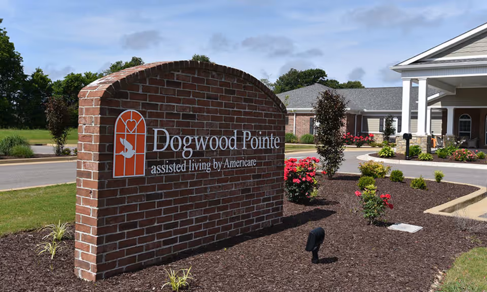 Brick sign for Dogwood Pointe assisted living by Americare in front of a landscaped area with flowers and shrubs, with part of the senior living facility building visible in the background under a partly cloudy sky.