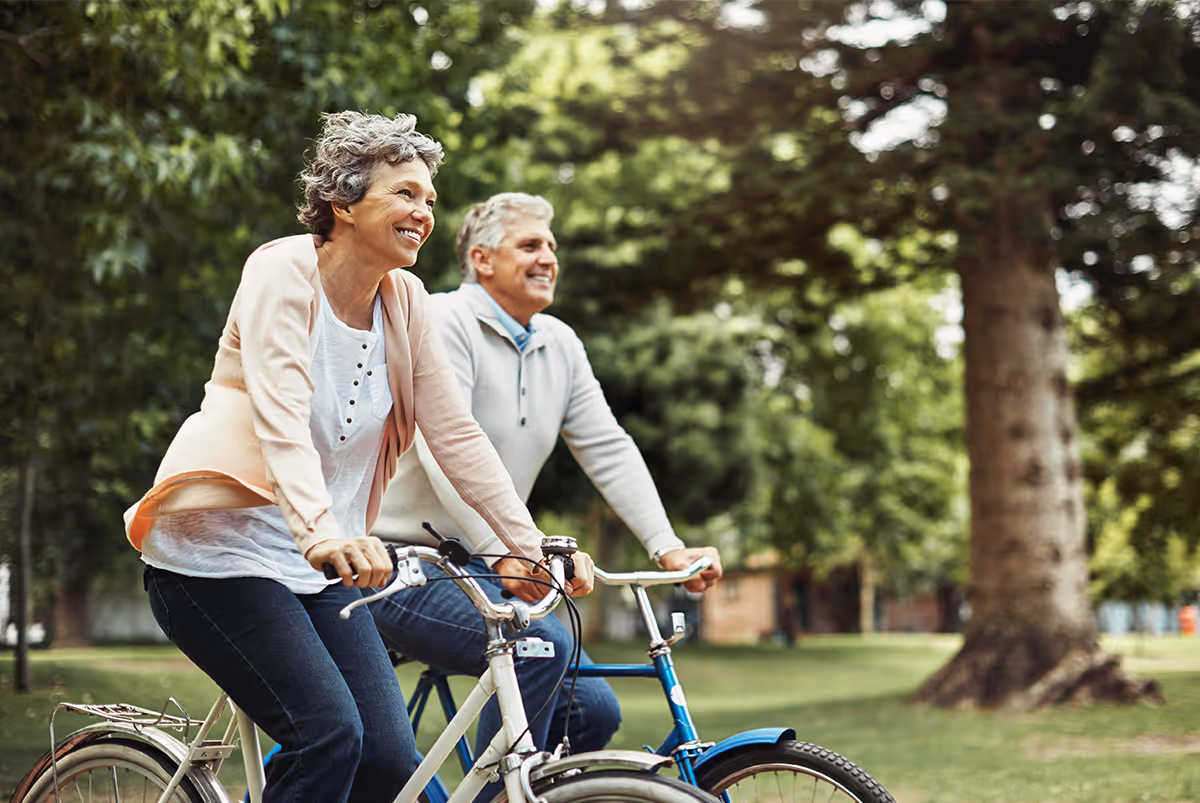 An older woman and man riding bicycles outdoors in a park-like setting with green trees and grass, both smiling and enjoying the activity.