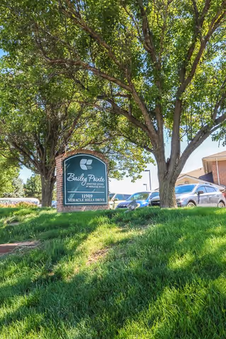 Outdoor view of a green grassy area with large leafy trees and a brick sign that reads 'Bailey Pointe Assisted Living at Miracle Hills, 11909 Miracle Hills Drive' with parked cars and a building partially visible in the background.