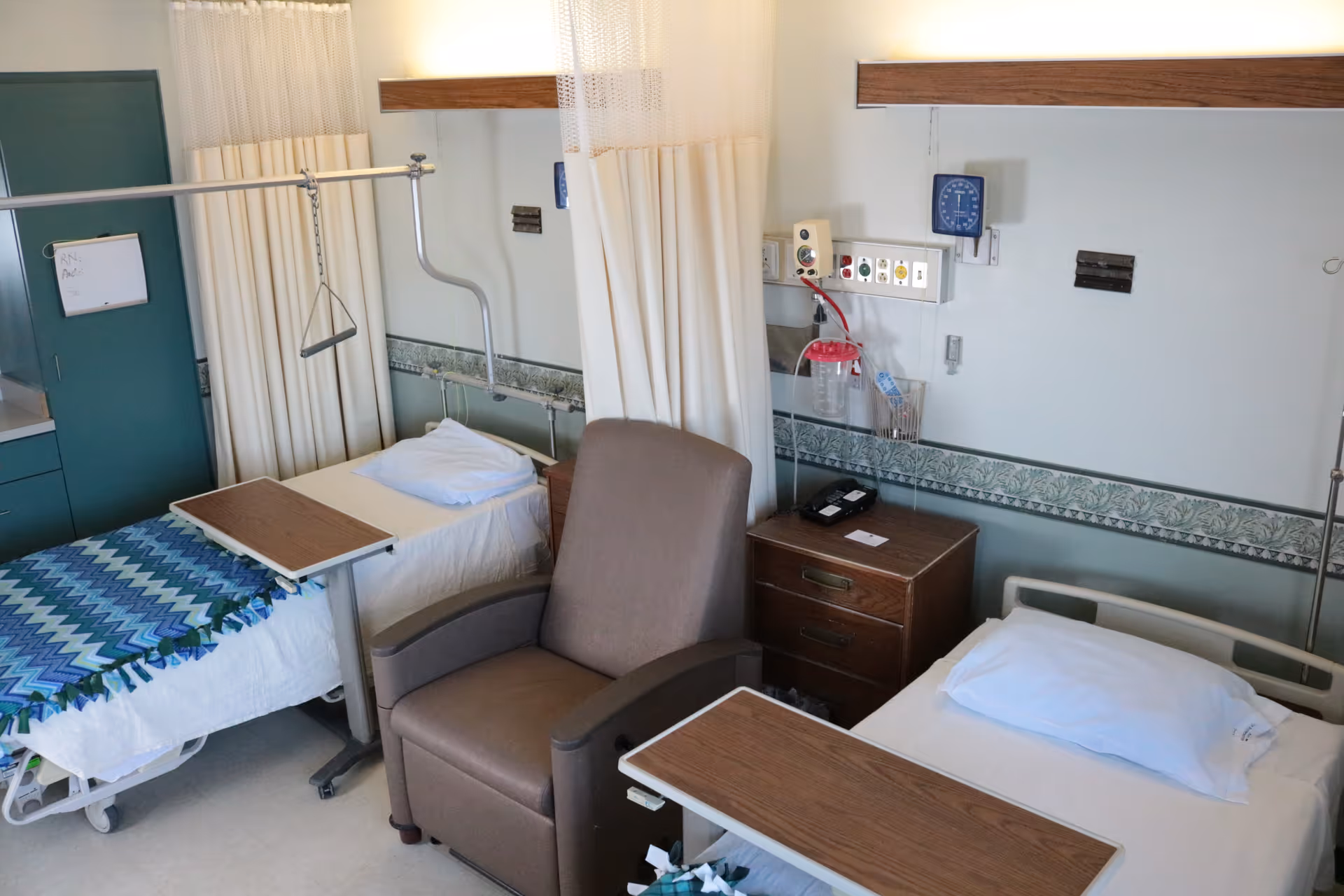 Two hospital beds separated by a privacy curtain with a recliner, bedside tables, over-bed trays and wall-mounted medical equipment in a patient room.