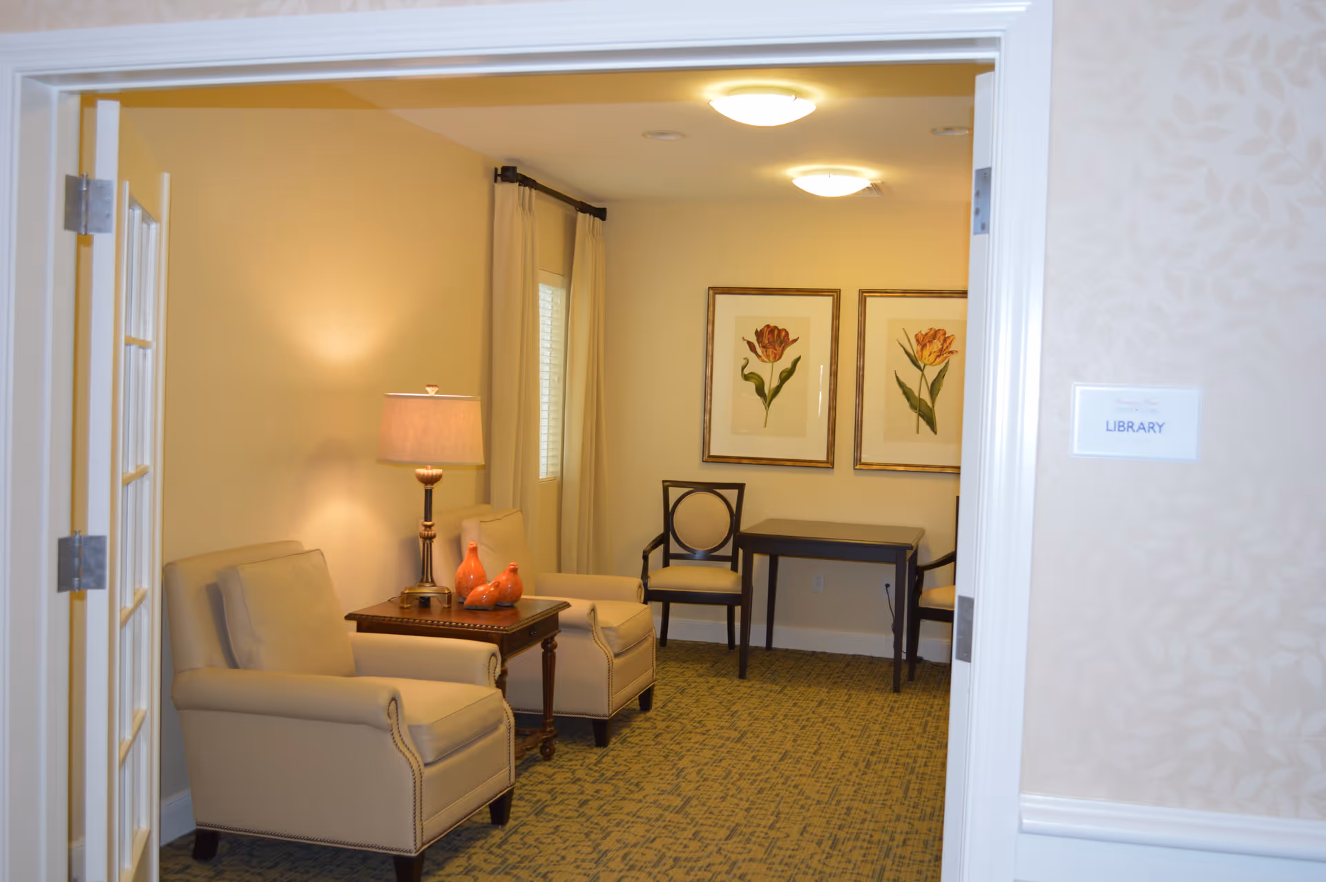 A cozy room labeled as a library with two beige armchairs, a wooden side table with a lamp and decorative items, two framed floral prints on the wall, a small table with two chairs, and beige walls with a patterned carpet.