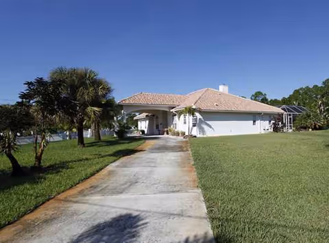 A single-story white building with a tile roof, driveway, palm trees, and green lawn.