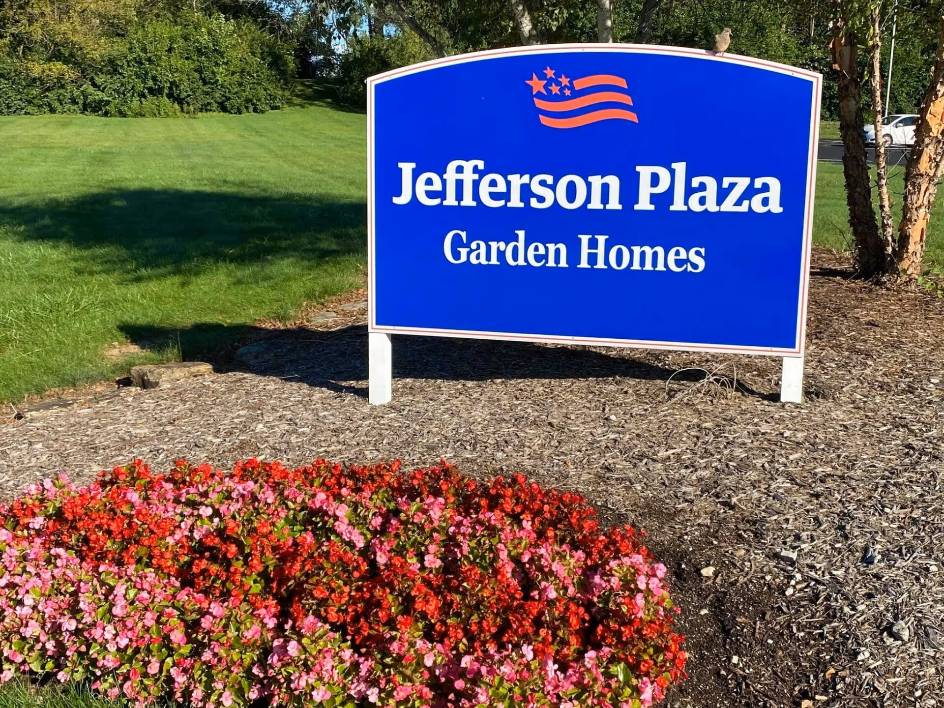 Blue sign reading 'Jefferson Plaza Garden Homes' in a landscaped area with pink and red flowers and a green lawn.