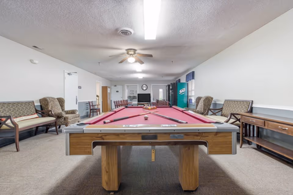 A recreational room with a red felt pool table in the center, pool cues and balls arranged on it. The room has several chairs and benches along the walls, a ceiling fan with light, fluorescent ceiling lights, a vending machine, a TV, and a clock on the far wall.