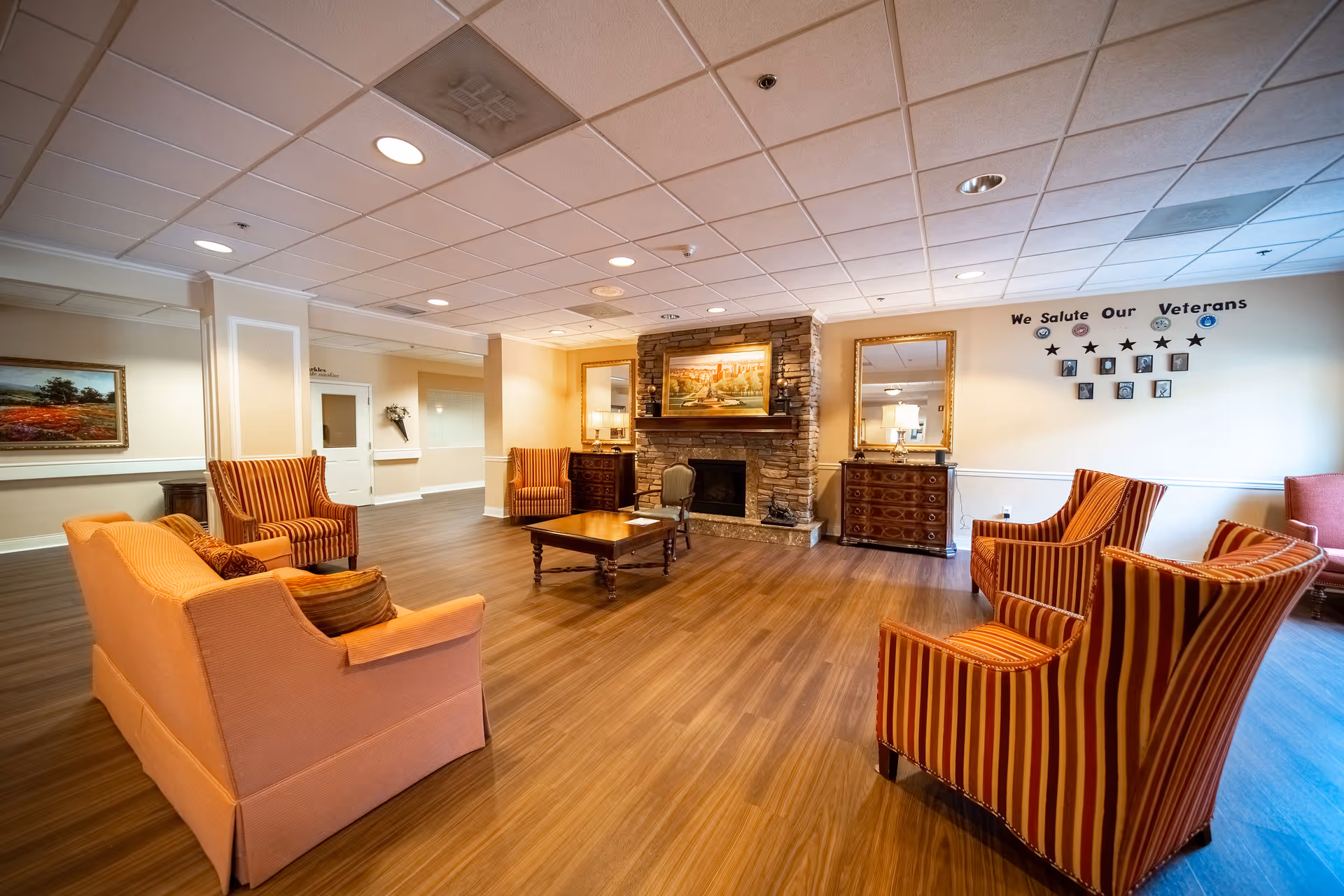 A spacious and warmly lit living room area with wooden flooring, featuring a peach-colored sofa, several striped armchairs, a wooden coffee table, and a stone fireplace with a painting above it. The walls are decorated with mirrors, a painting, and a tribute display for veterans. The ceiling has recessed lighting.