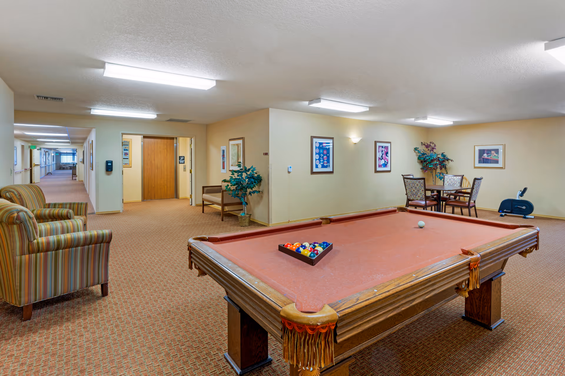 Community lounge with a pool table, seating area, and a small table and chairs in a carpeted senior living facility.