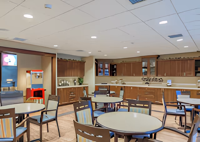 A spacious dining area with several round tables and chairs arranged around them. The room features a kitchenette with wooden cabinets, a countertop, and a sink along the back wall. There is a popcorn machine and a refrigerator visible on the left side of the room. The ceiling has recessed lighting, and the floor is carpeted.