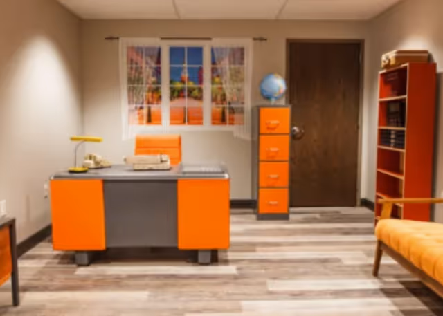 Office-style interior with an orange desk, matching filing cabinet and shelving, and an orange upholstered seat beneath a window.