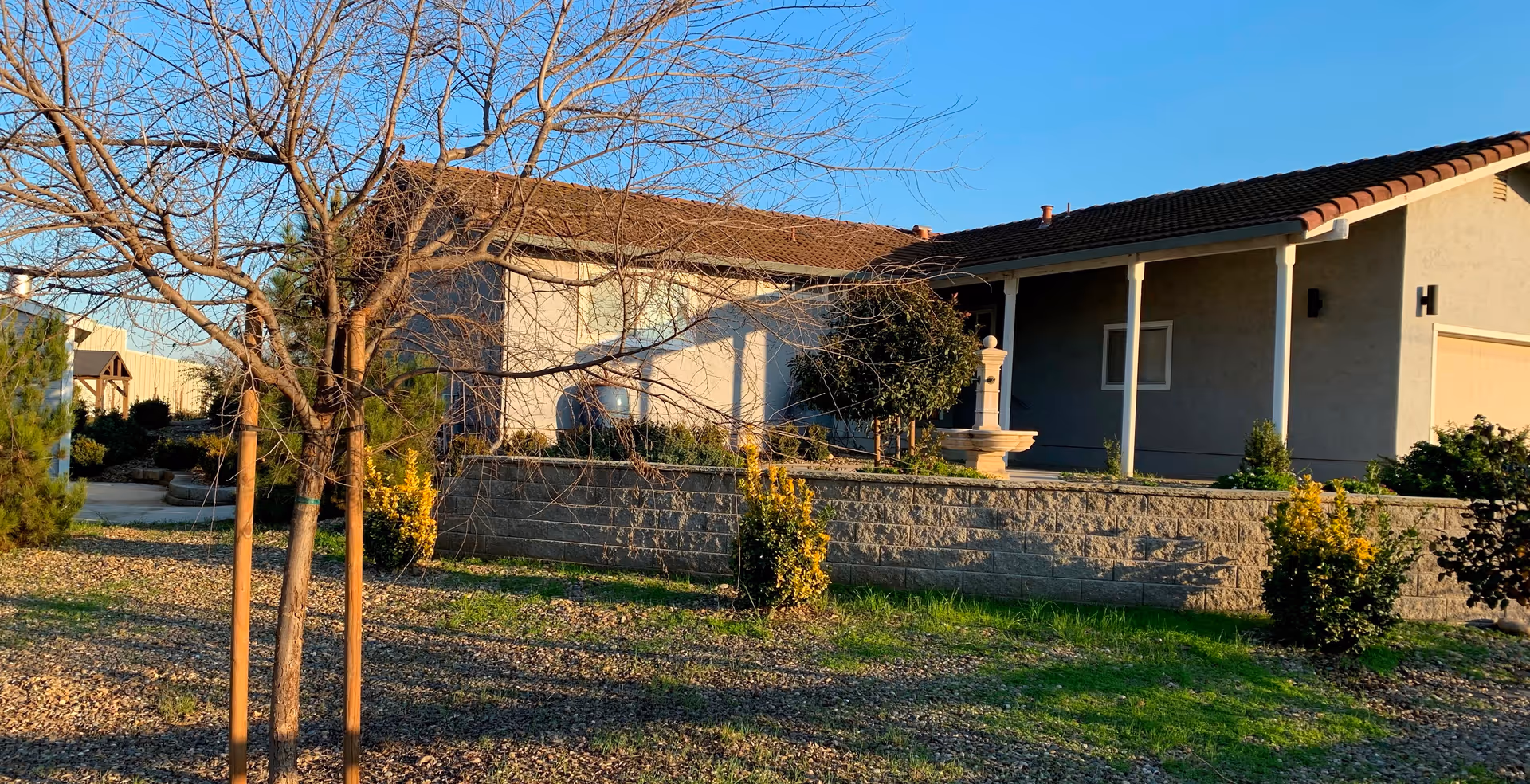 Single-story stucco building with a tiled roof, front porch, stone retaining wall, and trees in the yard.