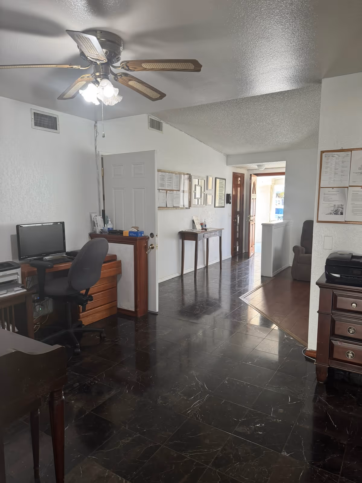 Interior view of a senior living facility office area with a ceiling fan, a computer desk with a chair, a small table along the hallway with framed documents on the wall, and a partially visible armchair in an adjacent room.