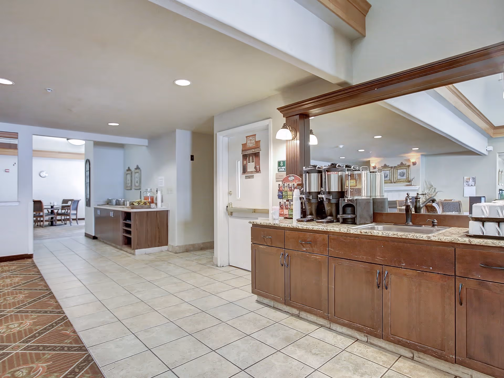 Interior view of a senior living facility showing a beverage station with multiple coffee dispensers on a granite countertop with wooden cabinets below. There is a large mirror behind the counter reflecting part of the room. To the left, there is a hallway leading to a dining area with tables and chairs. The floor is tiled and the ceiling has recessed lighting.