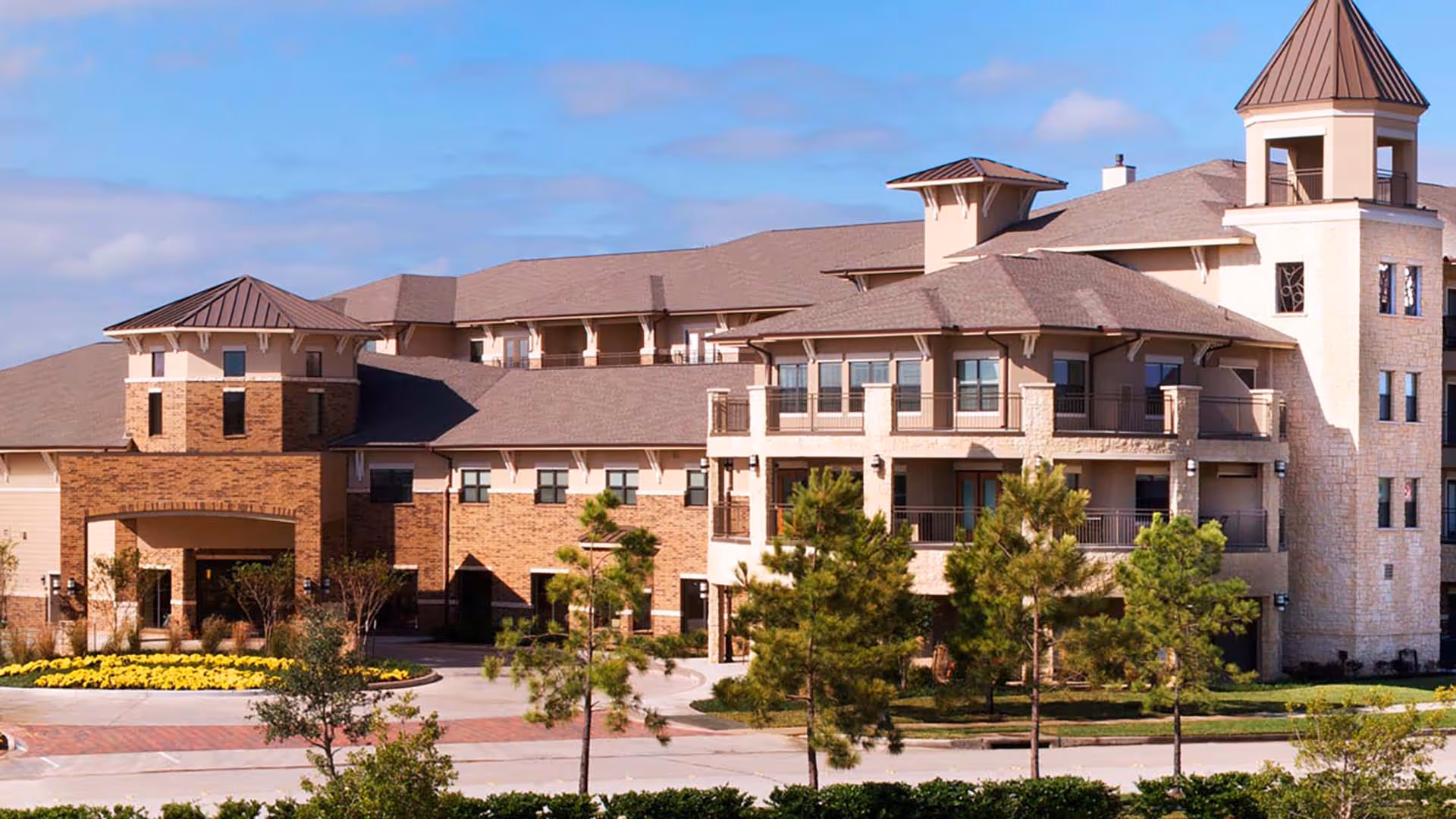 Exterior view of a large senior living facility building with multiple stories, a combination of brick and stone facade, several windows, balconies, and a driveway with landscaped greenery and small trees in front.