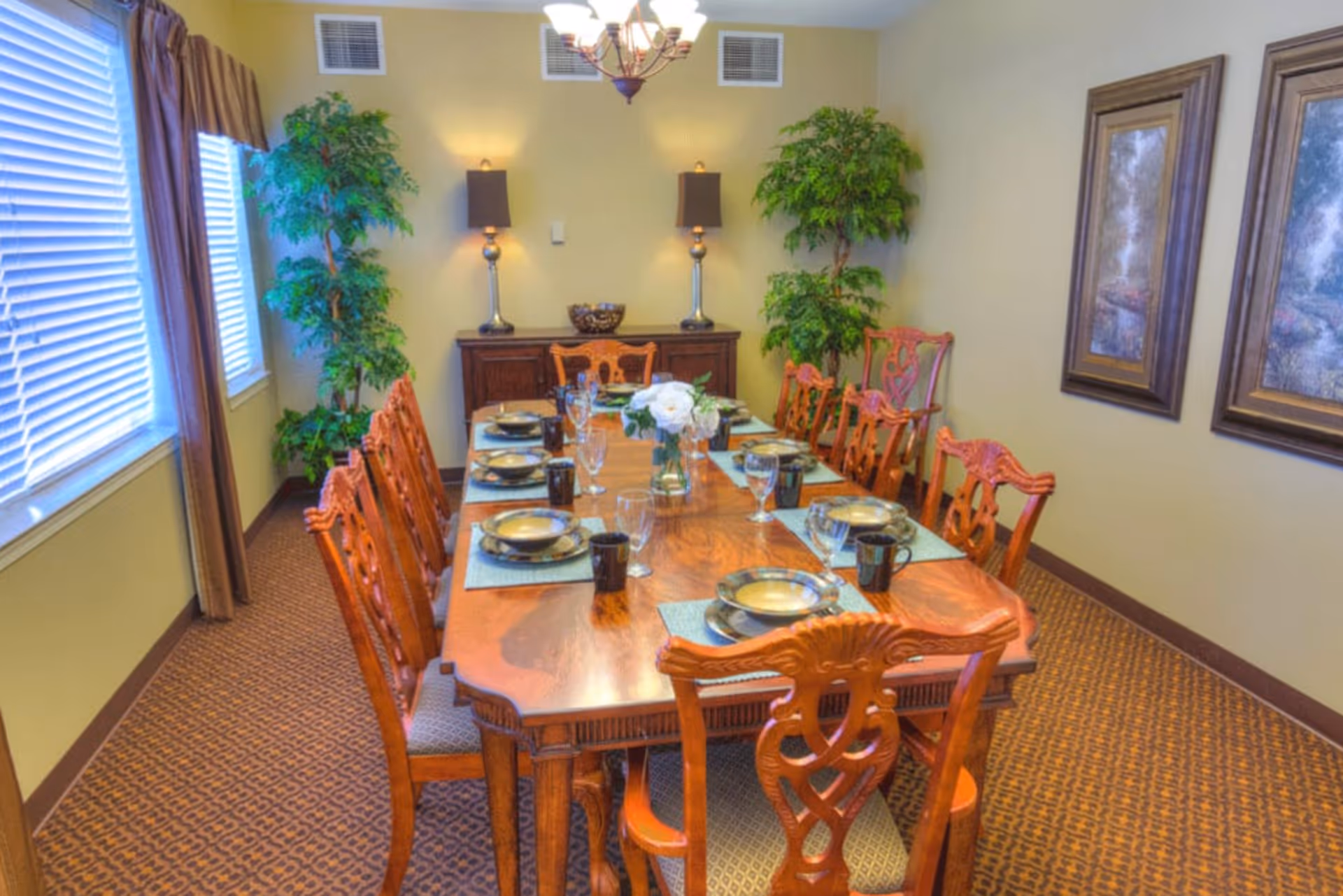 A formal dining room with a long wooden table set for eight people. The table has plates, bowls, glasses, and mugs on placemats. There are wooden chairs with intricate backs around the table. The room has two large windows with blinds and curtains on the left side, two framed paintings on the right wall, two tall green plants in the corners, a sideboard with two lamps and a decorative bowl against the back wall, and a chandelier hanging from the ceiling.