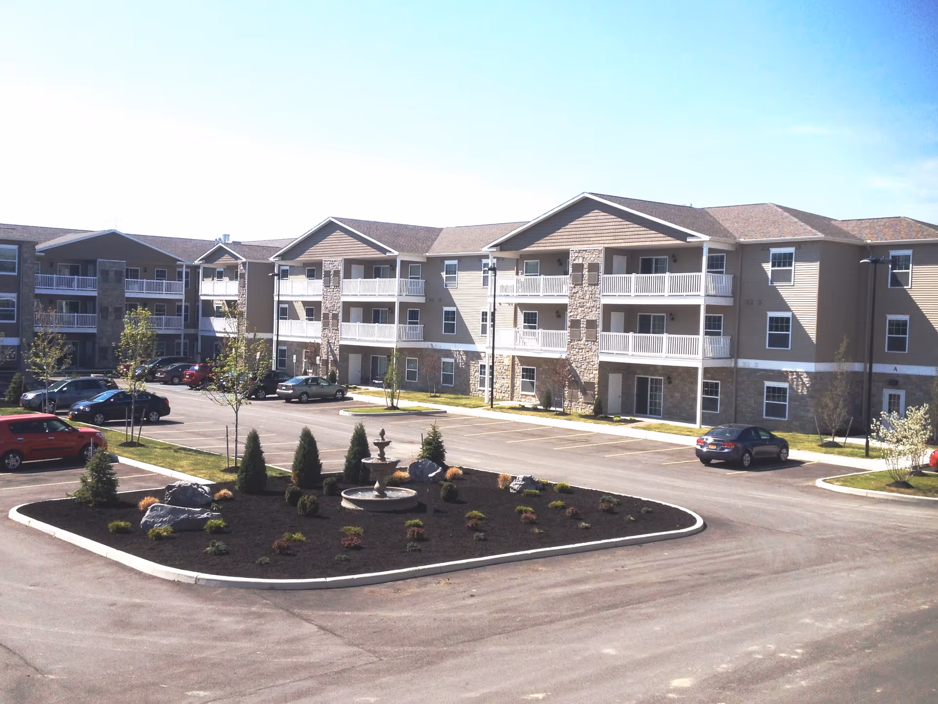 Exterior view of a multi-story residential building with balconies, surrounded by a parking lot and landscaped area with small trees, shrubs, rocks, and a tiered water fountain in the center.