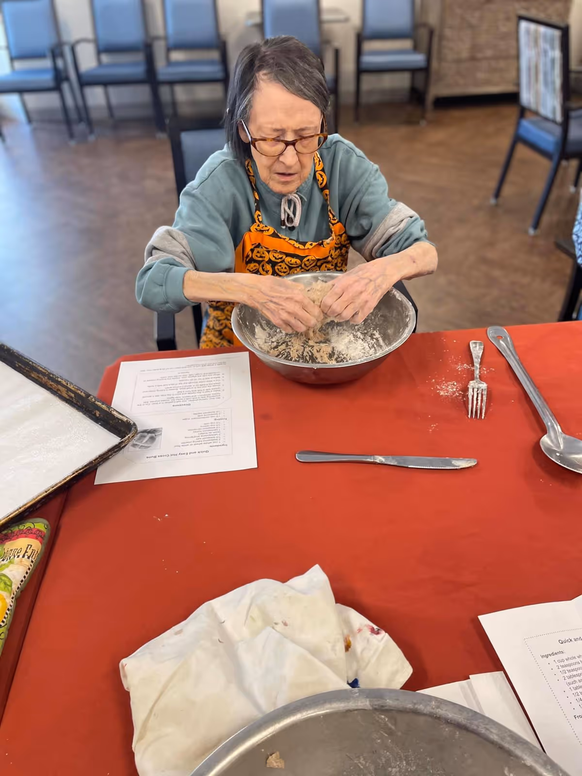 An elderly woman wearing glasses and a pumpkin-themed apron is sitting at a table kneading dough in a large metal bowl. The table is covered with a red tablecloth and has various utensils, a baking sheet, and printed recipe sheets on it. The background shows a room with several empty chairs.