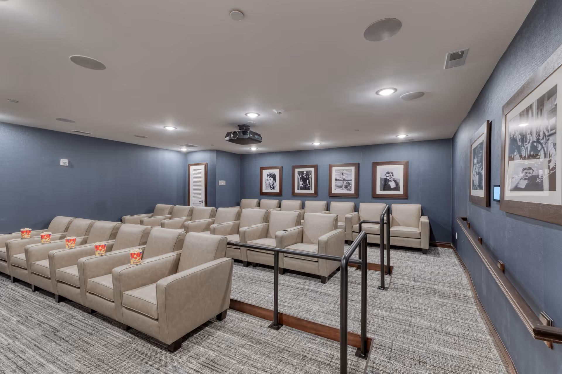 A small theater room with three rows of beige armchairs arranged on carpeted tiered flooring. Each chair has a popcorn container placed on the armrest. The walls are painted blue and decorated with framed black and white photographs. A ceiling-mounted projector is visible above the seating area.