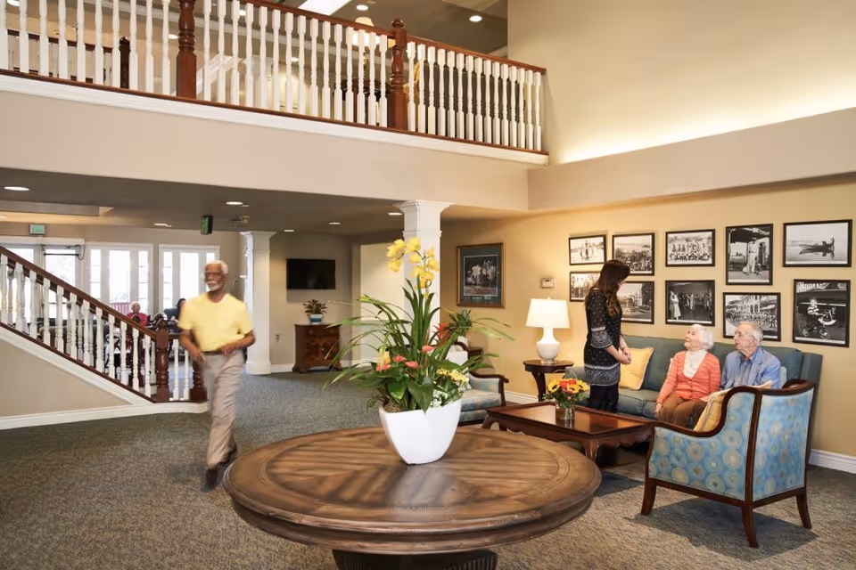 A spacious and well-lit senior living facility common area with a round wooden table holding a large white planter with yellow and red flowers in the foreground. To the right, a woman is standing and talking to two elderly people seated on a blue couch and chair. The walls are decorated with framed black and white photographs. In the background, a man in a yellow shirt is walking near a staircase with white railings and wooden banisters. The area has carpeted floors and a second-floor balcony with white railings.
