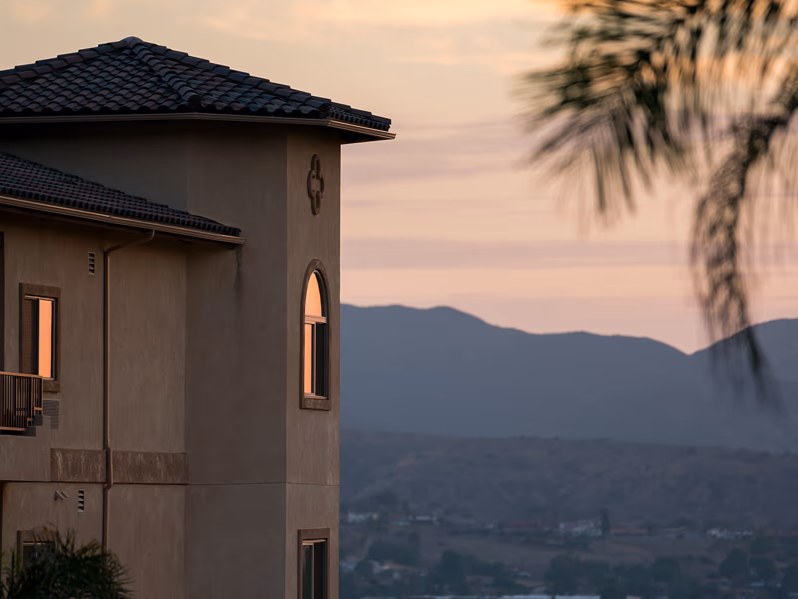 Side view of a senior living facility building with a tiled roof and arched windows at sunset, with mountains and a valley in the background and a blurred palm tree in the foreground.
