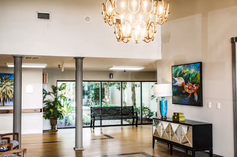 Bright lobby-style common area with hardwood floors, columns, a decorative chandelier, bench by large windows, and a console table with lamp and artwork.