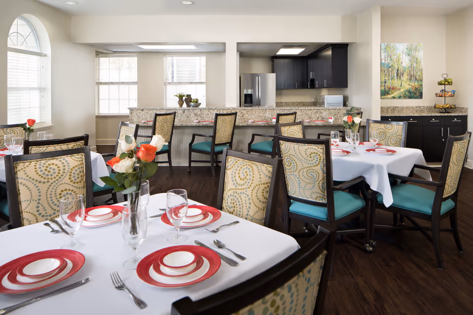A bright dining room with several tables set with white tablecloths, red and white plates, silverware, and glassware. Each table has a vase with red and white roses. The room features patterned chairs with teal cushions, large windows with blinds, and a kitchen area in the background with dark cabinets, a refrigerator, and a granite countertop. A colorful painting and a tiered fruit basket are visible on the right side.