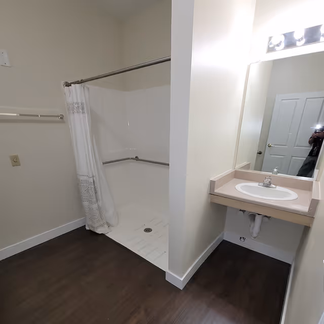 A bathroom with a walk-in shower featuring a white curtain and grab bars, a sink with a countertop and a large mirror above it, and a wooden floor.