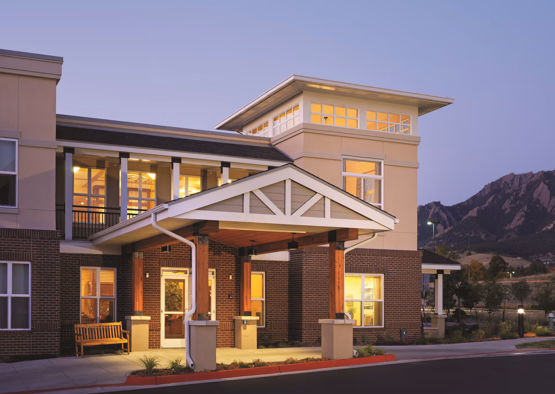Exterior view of Flatirons Terrace Senior Living building at dusk, showing the entrance with a covered porch supported by wooden beams, illuminated windows, and a bench near the door. Mountains are visible in the background under a clear sky.