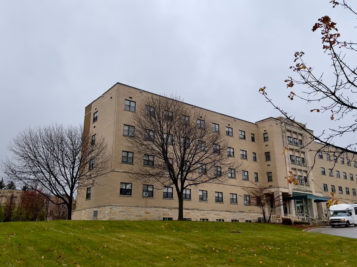 Five-story beige brick senior living building with leafless trees and a grassy lawn under a cloudy sky.
