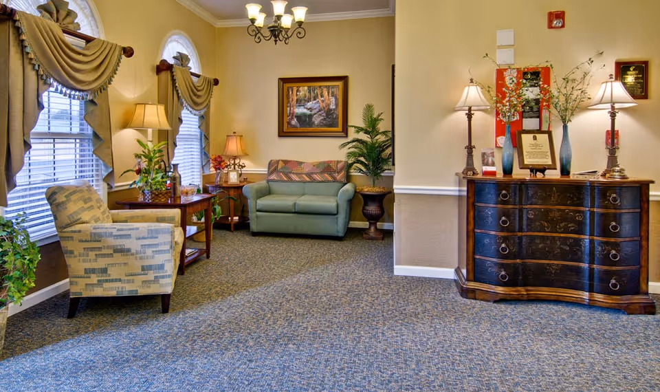 A cozy living room area in a senior living facility with patterned carpet, two windows with draped curtains, a patterned armchair, a small wooden side table with a lamp and plants, a green loveseat with a decorative pillow, a framed nature painting on the wall, a large potted plant, and a dark wooden chest of drawers with two table lamps and decorative vases on top.