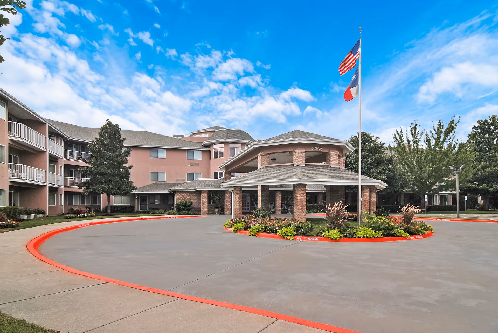 Front entrance of a multi-story senior living building with a covered porte-cochère, circular driveway, landscaped island and a flagpole flying the U.S. and Texas flags.