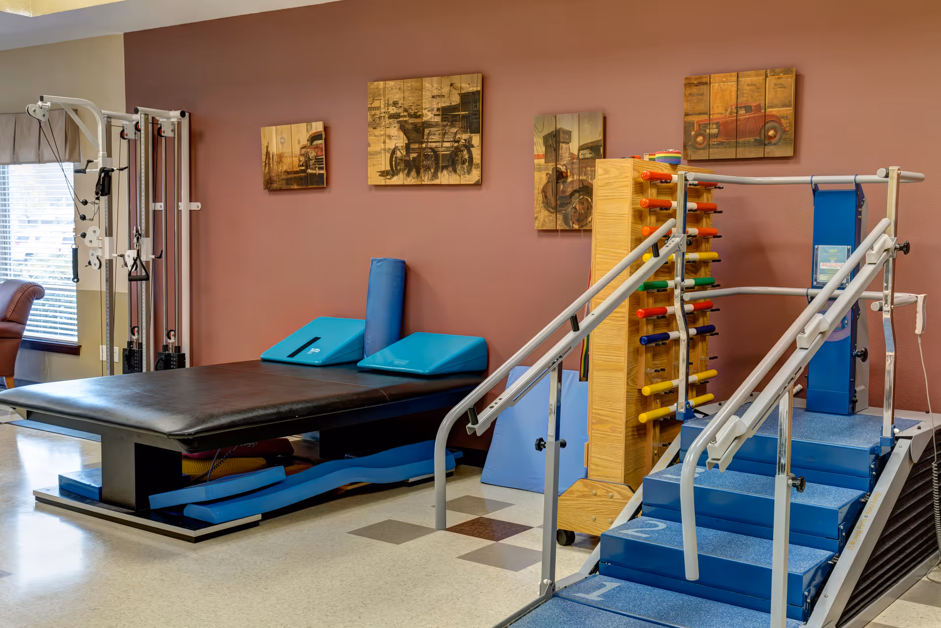Physical therapy room with exercise equipment including a padded therapy table, blue foam wedges, a set of stairs with handrails for rehabilitation, and a rack holding colorful exercise bands. The room has a brown wall with vintage vehicle artwork and a window with blinds letting in natural light.