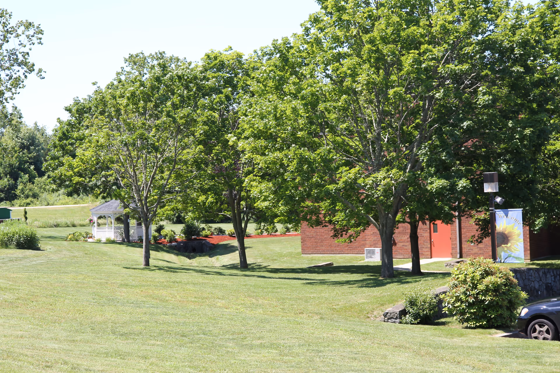 A grassy outdoor area with several green leafy trees, a small white gazebo in the background, a brick building partially visible on the right side, and a parked car near some bushes.