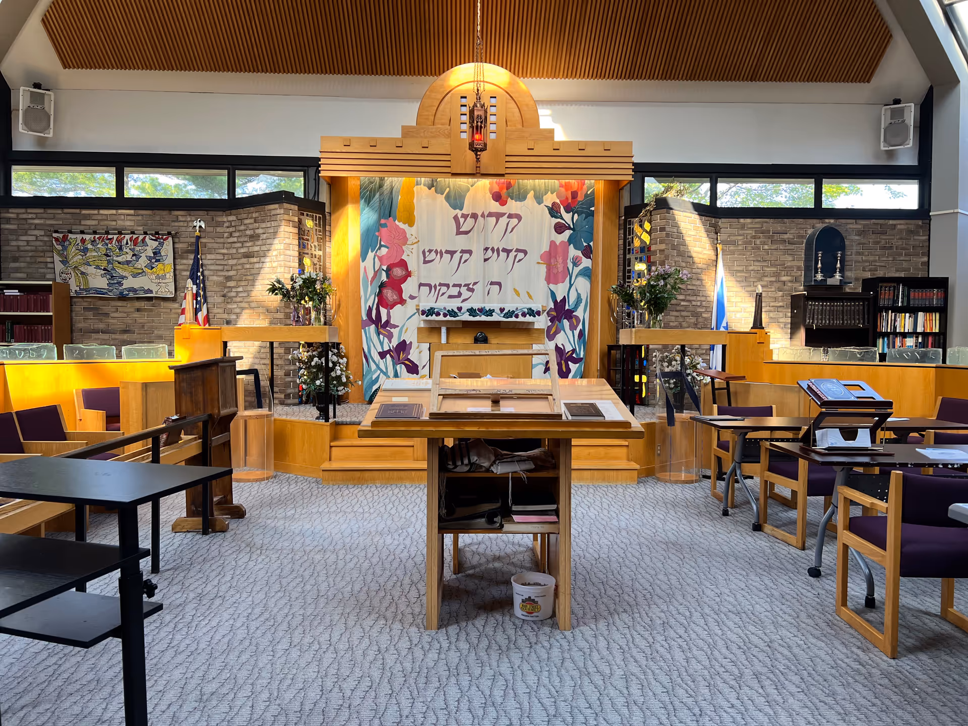 Interior view of a synagogue or chapel with wooden furniture, a central podium, and a decorative curtain with Hebrew text and floral designs behind the altar. The room has carpeted floors, large windows near the ceiling, and bookshelves along the walls.