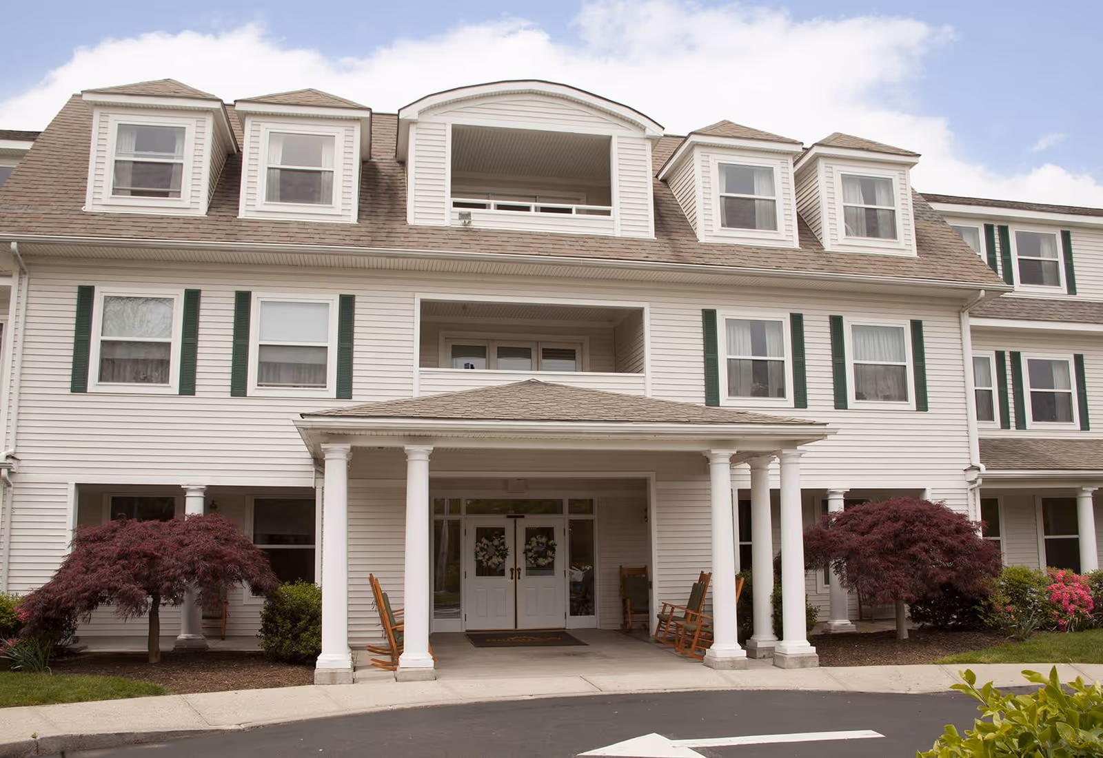 Front entrance of a multi-story white senior living building with columns, rocking chairs on the porch, and landscaping.