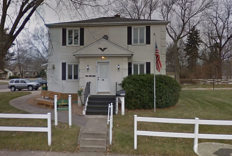 Front exterior view of a two-story white building with black shutters, a small porch with steps leading up to a white door, an American flag on a flagpole to the right, a green bush, a white fence in the foreground, and a bench on the left side near a curved sidewalk.