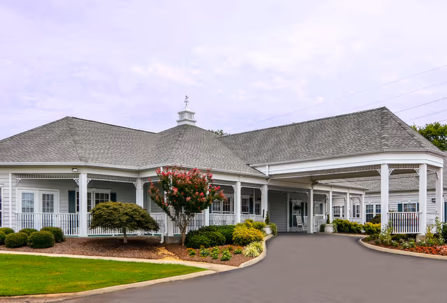 Exterior view of The Neighborhood at Sheffield facility showing a single-story building with a covered entrance, white railings, and landscaped bushes and trees in front.