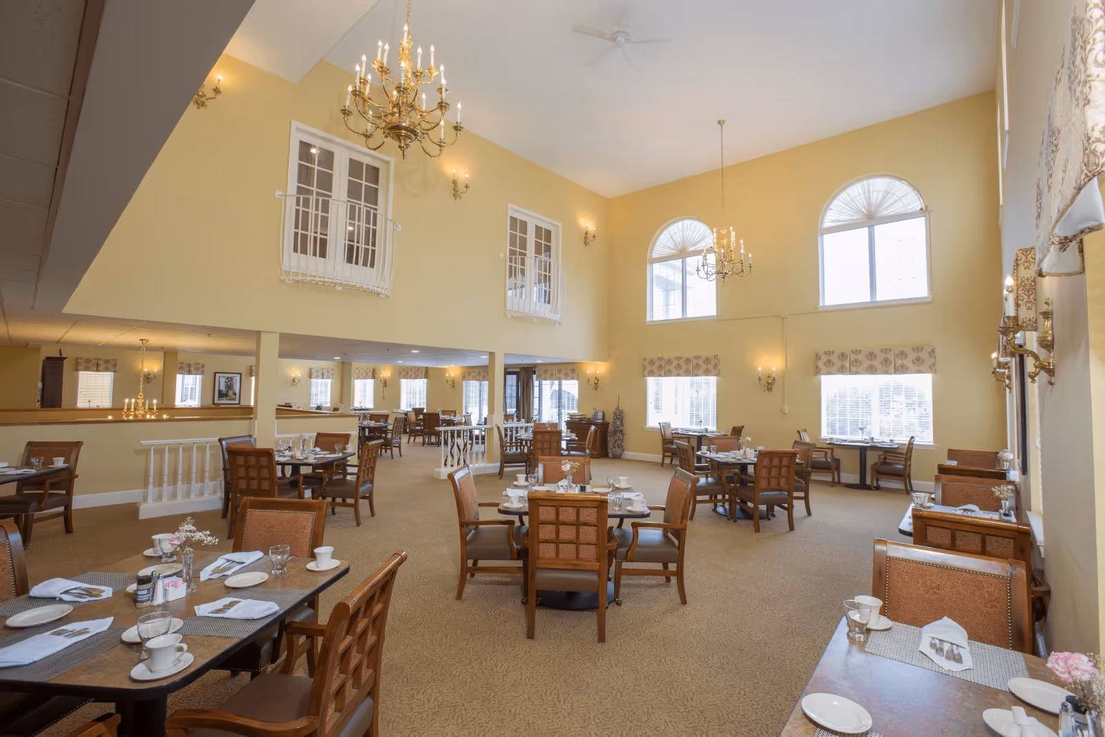 A spacious dining room with high ceilings and large arched windows letting in natural light. The room is furnished with multiple wooden tables and chairs, each set with white plates, cups, and napkins. Elegant chandeliers and wall sconces provide additional lighting. The walls are painted a soft yellow, and the carpet is beige with a subtle pattern.