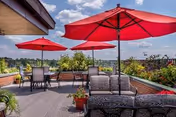 Outdoor patio area with multiple red umbrellas providing shade over tables and chairs. There are plants and flowers in planters along the edge of the patio, with a clear blue sky and some clouds in the background.