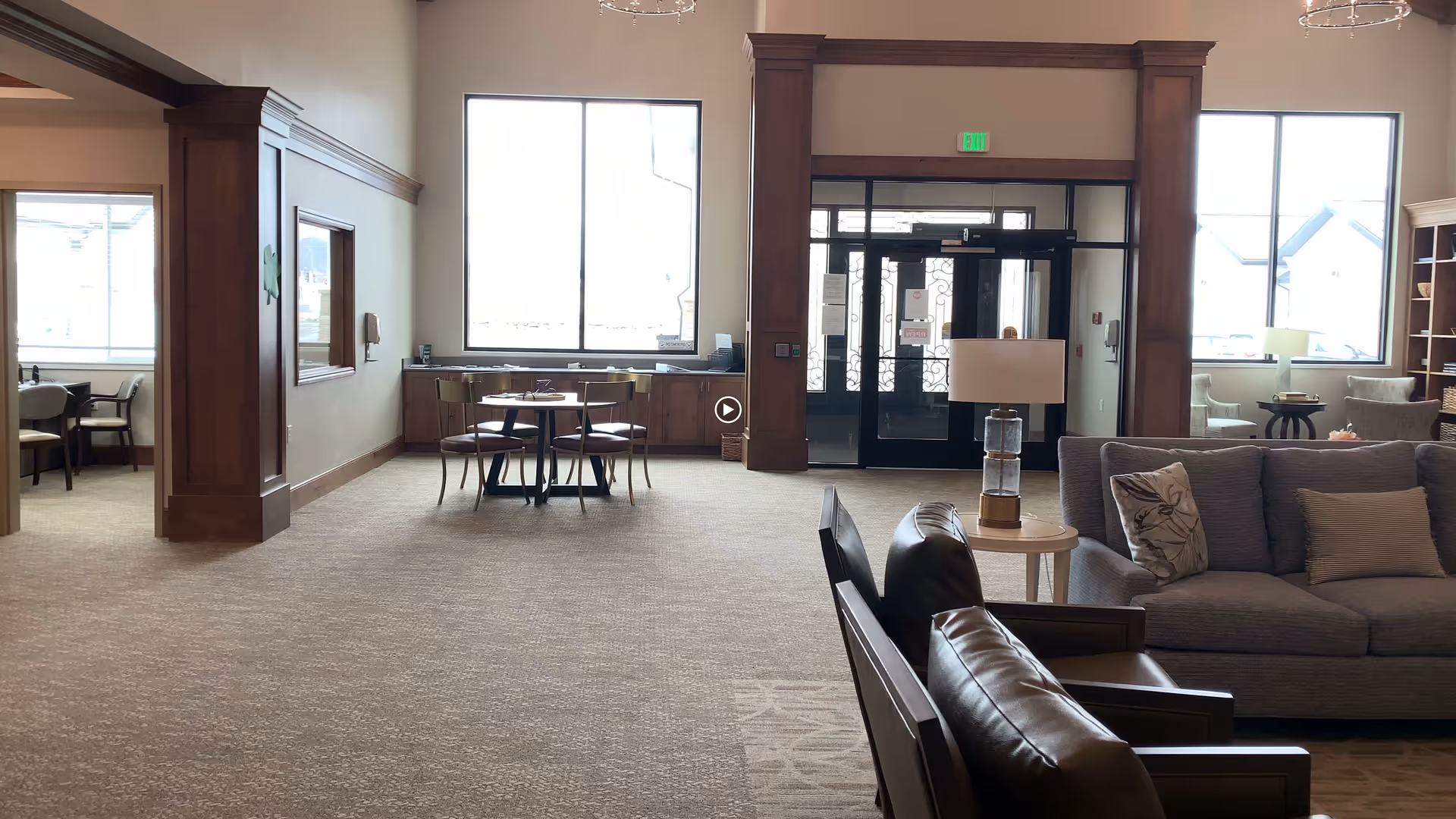 Spacious lobby area with sofas and armchairs, a small round dining table, large windows, and double glass entrance doors.