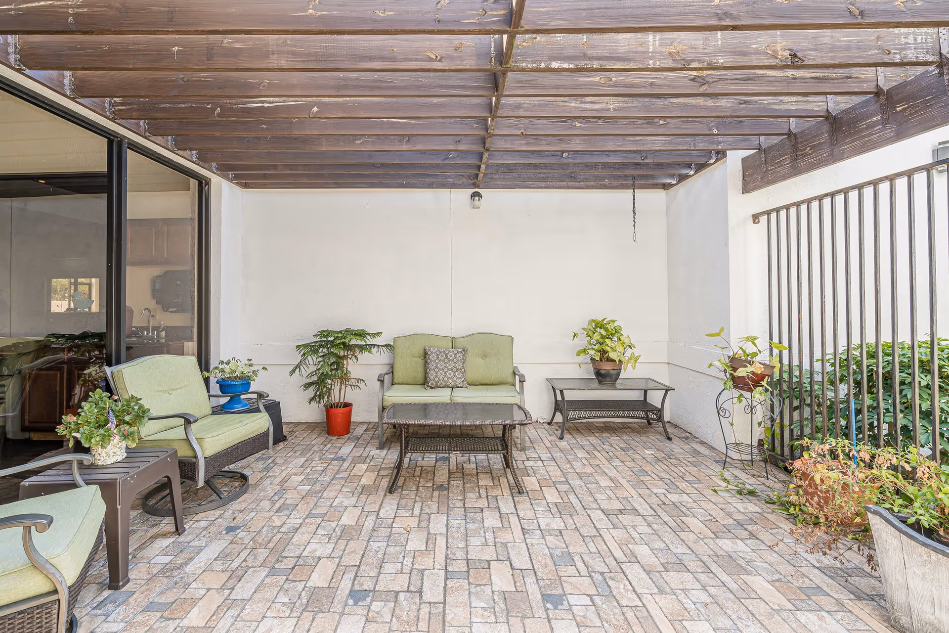 Covered outdoor patio area with brick flooring, green cushioned seating including a loveseat and chairs, a glass-top coffee table, and various potted plants. The patio has a wooden pergola overhead and is adjacent to a building with large glass windows.