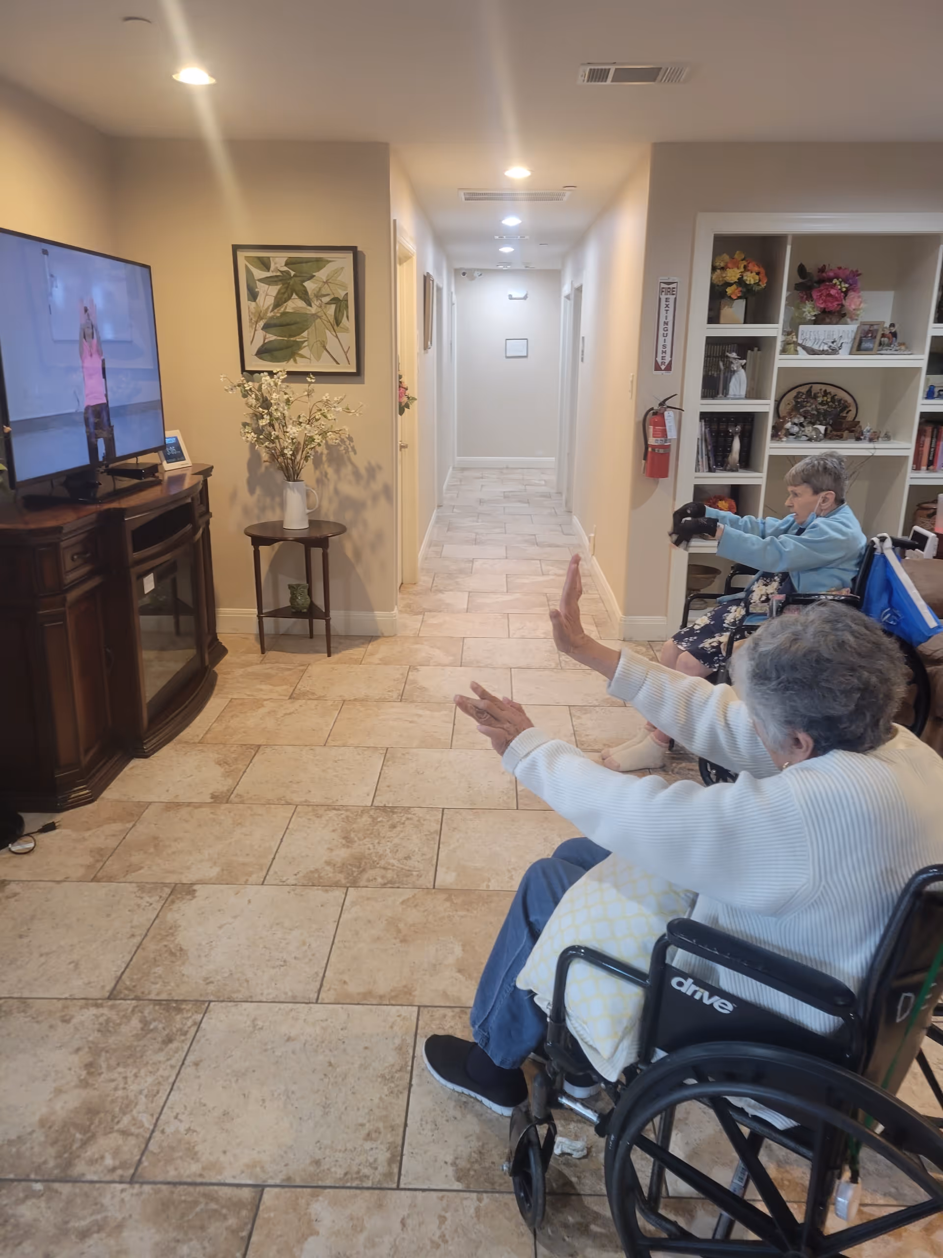 Two elderly women in wheelchairs participating in a seated exercise session while watching a fitness instructor on a television screen in a hallway of an assisted living facility. The hallway has tiled floors, beige walls, and decorative shelves with flowers and framed pictures.