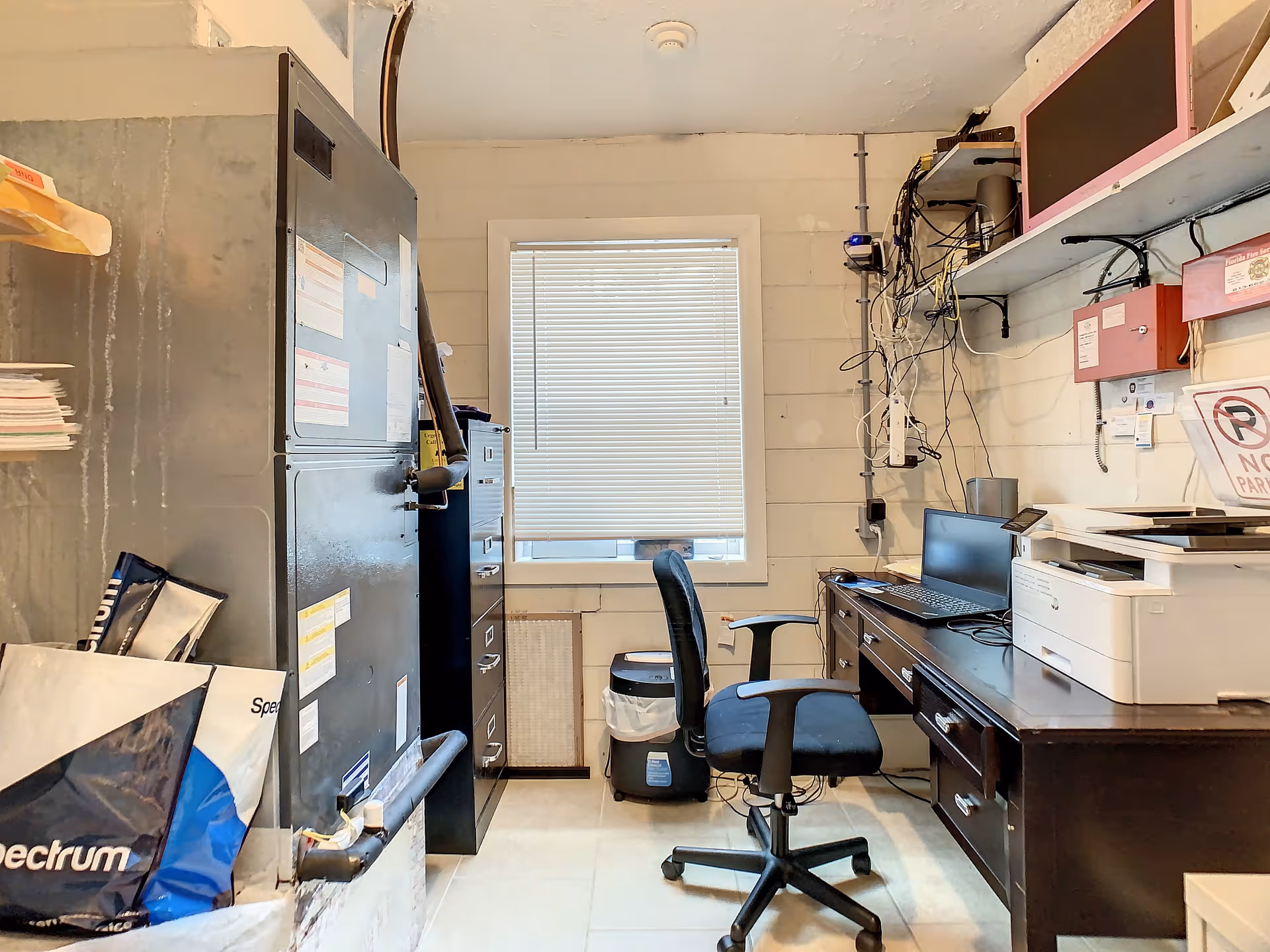 Small cluttered office/storage room with a desk, office chair, filing cabinets, printer and exposed cables beneath a window.