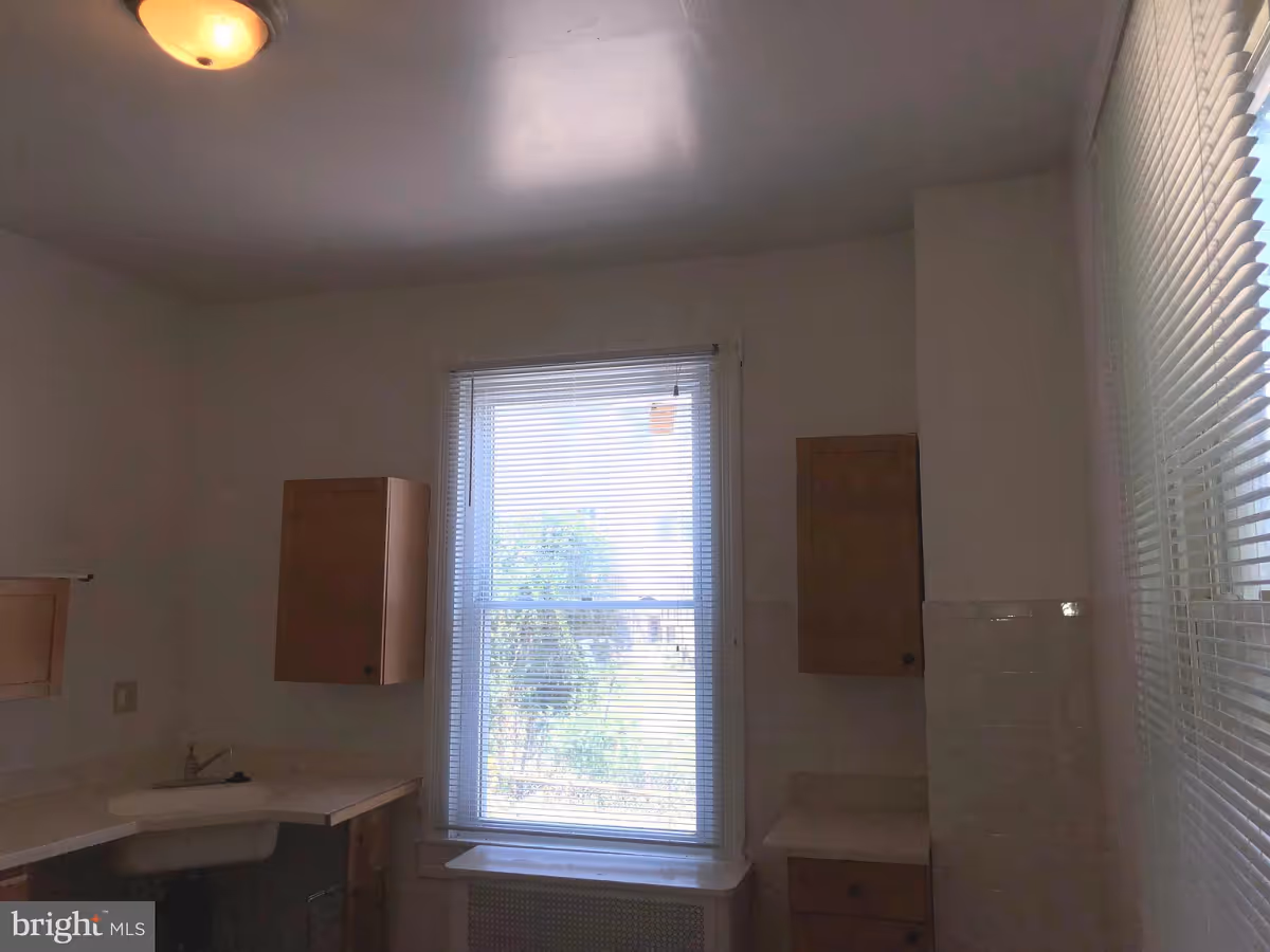 Interior view of a small kitchen area with a window in the center covered by blinds. There are wooden cabinets mounted on the walls and a countertop with a sink below the window. The ceiling light is on, and there is another window with blinds on the right side.