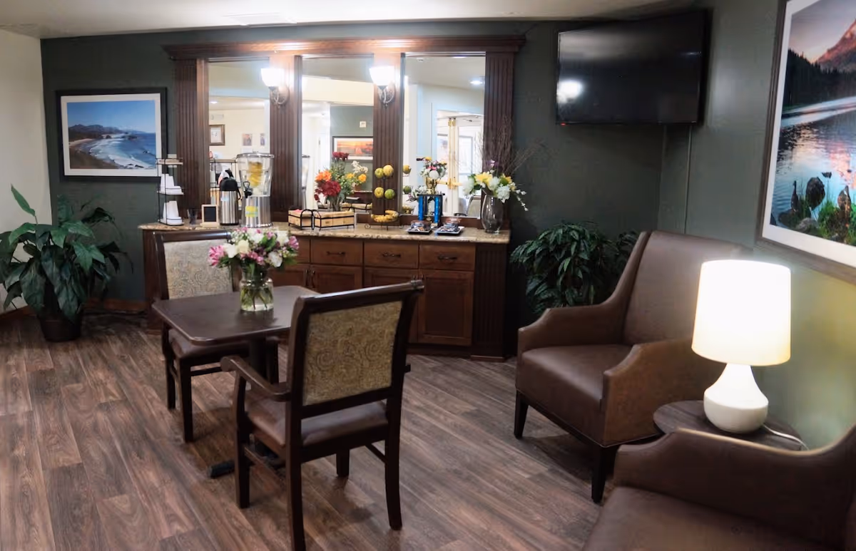 A cozy senior living lounge with chairs, a small table with flowers, a buffet with beverages and decor, and a wall-mounted TV.