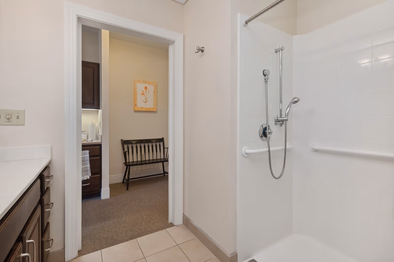 Bathroom with a white tiled walk-in shower equipped with grab bars and a handheld showerhead. To the left, there is a countertop with dark wood cabinets and drawers. Through the doorway, a small seating area with a black wooden bench and a framed floral artwork on the wall is visible.